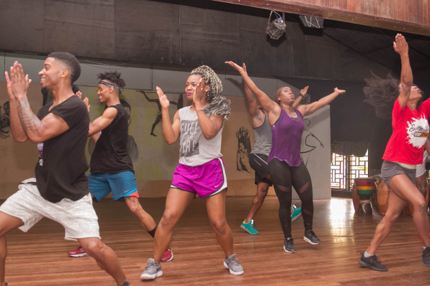 Eine Gruppe von Menschen tanzt in einem Zumba-Kurs in einem Studio mit einem Gemälde an der Wand, einem Glasfenster rechts und Deckenlampen.