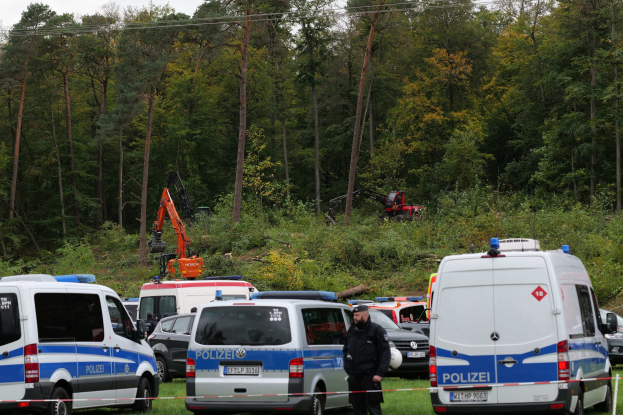 Polizeiautos auf einem Parkplatz mit einer Person in der Nähe, Bäume, Pflanzen, ein Bagger und Himmel im Hintergrund.