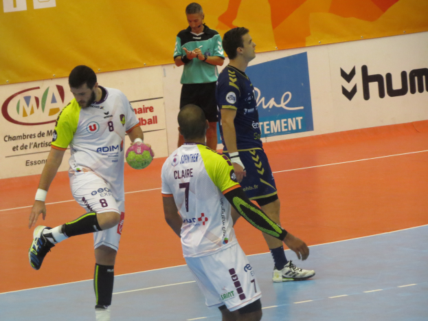 Eine Gruppe von Männern, die Handball auf einem Feld spielen, wobei einer einen Ball hält und im Hintergrund Schilder mit Text zu sehen sind.