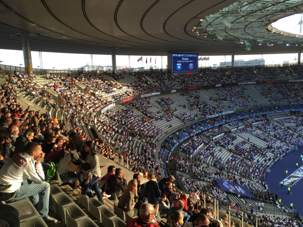 Eine große Menschenmenge sitzt im Allianz Arena Stadion in München, Deutschland, und schaut ein Fußballspiel, mit einer Bühne auf der rechten Seite, Fahnen, Stangen und einem Bildschirm im Hintergrund, unter einem sichtbaren Himmel.