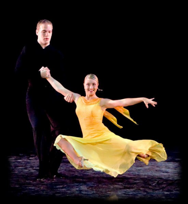 Ein Mann in einem schwarzen Anzug und eine Frau in einem hellen Kleid tanzen auf der Bühne, ihre Arme verschränkt und Gesichter strahlend vor Freude, während sie sich gegen einen dunklen blauen Hintergrund anschauen.