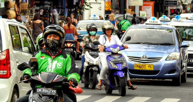 Eine Gruppe von Menschen auf Motorrädern fährt eine belebte Straße entlang mit Autos und anderen Fahrzeugen, einige Fahrer tragen Helme, Gebäude, Bäume, Schilder und Geschäfte im Hintergrund.
