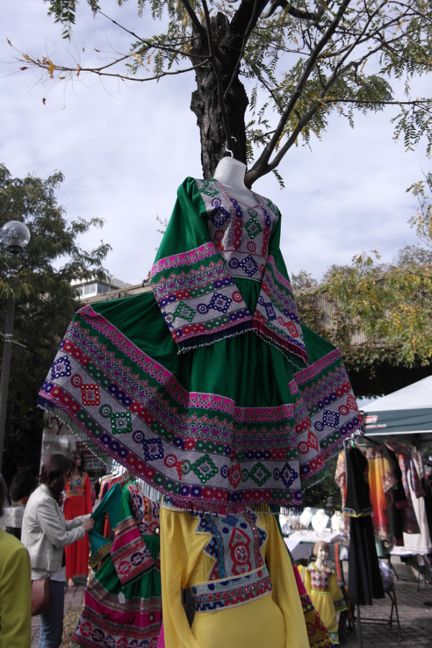Eine Schaufensterpuppe steht in der Mitte eines belebten Einkaufsgebiets mit verschiedenen Geschäften im Hintergrund, eine Person, die Kleider an einem Pfahl aufhängt, ein Gebäude und Bäume daneben.
