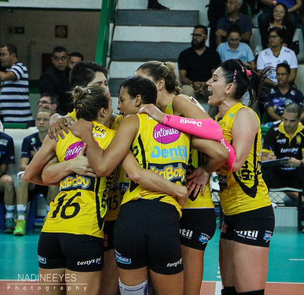 Eine Gruppe von Frauen-Volleyballspielerinnen in gelben T-Shirts und schwarzen Shorts auf einem Feld, mit Zuschauern und einer Wand im Hintergrund.