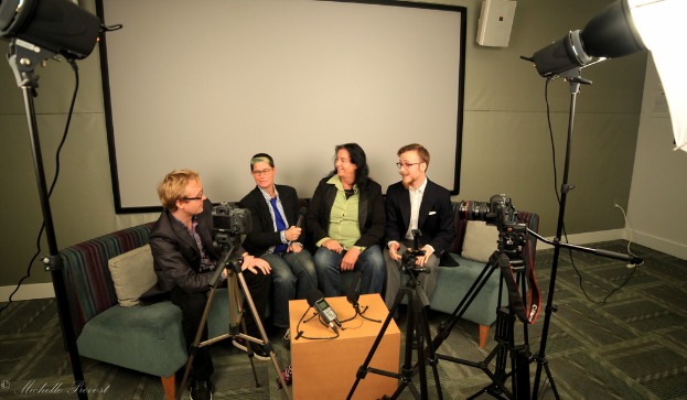 Eine Gruppe von drei Personen, die auf einer Couch sitzen und ein Interview geben, mit einer Person in schwarzer Jacke und Hose links, einer Person in einem Mantel und blauer Krawatte, die ein Mikrofon hält, in der Mitte, und einer Person, die rechts lächelt, mit einem Projektionsschirm dahinter, einem Mikrofonrecorder auf dem Tisch in der Mitte und Kameras mit Stativen auf beiden Seiten.