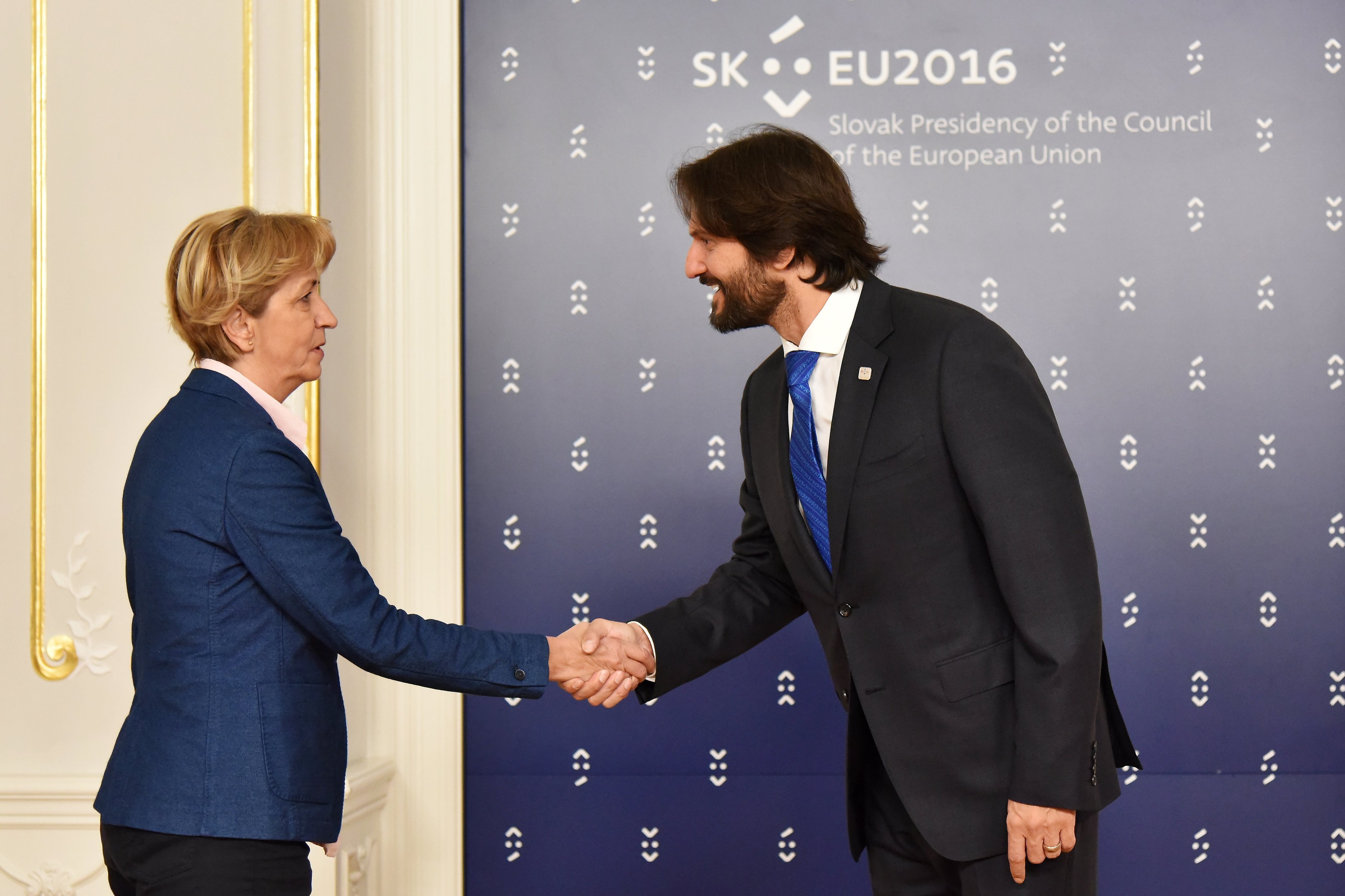 Bundeskanzlerin Angela Merkel und Bundeskanzlerin Joachim Gauck schütteln Hände vor einem 'EU-Gipfel'-Plakat, freundlich und einladend lächelnd.