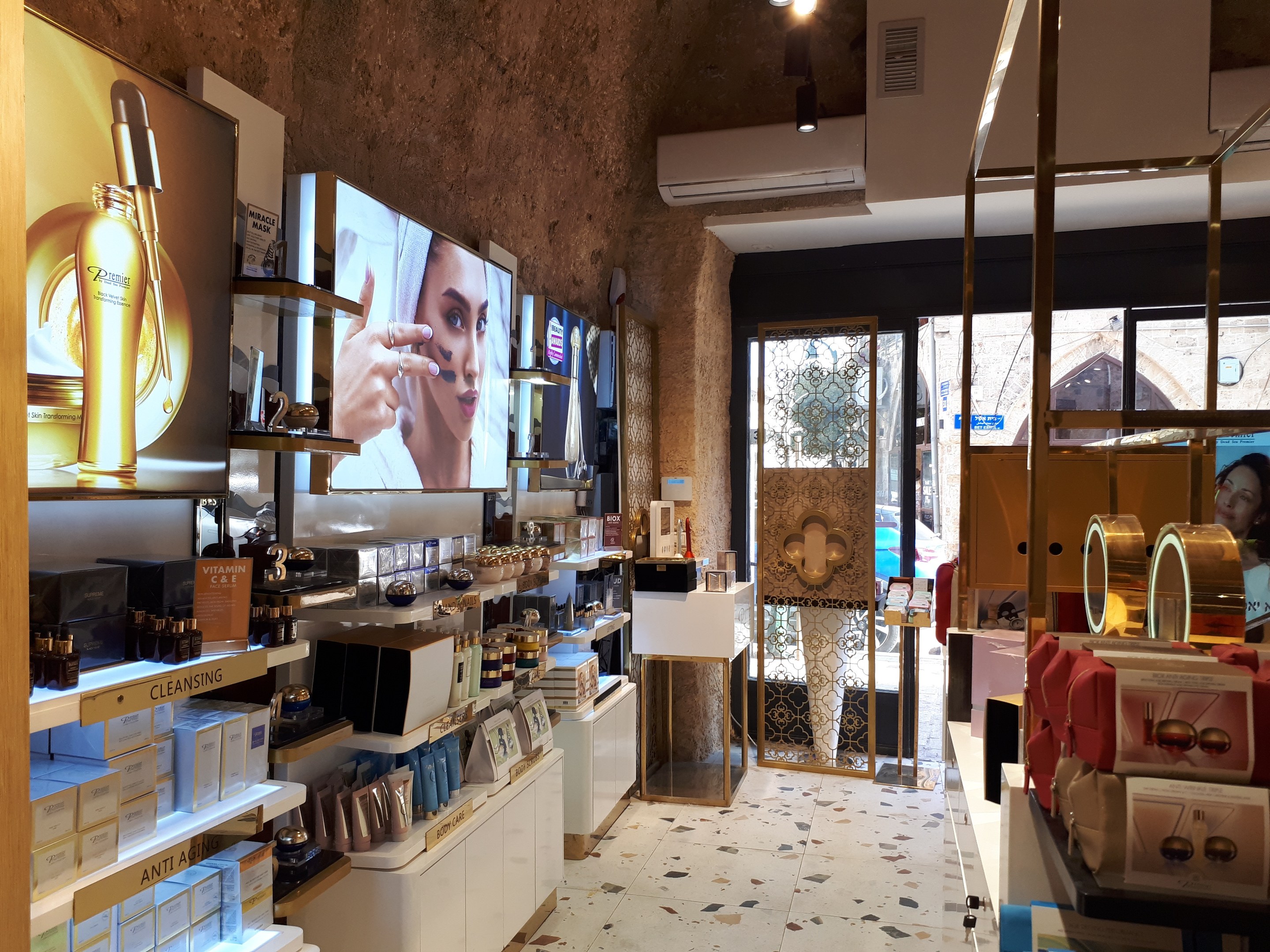 Ein Kosmetikgeschäft mit Regalen voller Flaschen, Schachteln und anderen Produkten, das Wandbildschirme, Deckenleuchten, eine Klimaanlage und Glastüren zeigt, durch die man eine Person, ein Auto und ein Gebäude sehen kann.