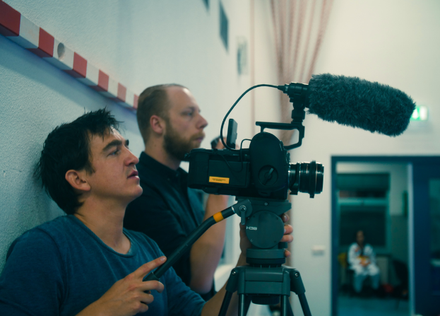 Ein Mann in einem blauen T-Shirt filmt mit einer Kamera, wobei ein Mann in einem schwarzen Hemd und eine Frau in einem weißen Kleid anwesend sind, mit einer weißen Wand und einem Vorhang im Hintergrund.
