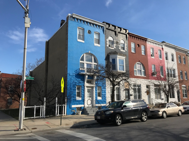Eine Reihe bunter Häuser an einer Straßenecke mit geparkten Autos, Bäumen, Strommasten und einem Zaun unter einem bewölkten Himmel in Baltimore, Maryland.