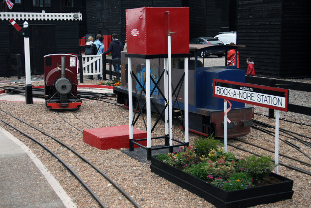 Spieldosen auf Schienen, ein Tank auf Ständern, Pflanzen, ein Schild, ein Mast mit einer Flagge, Menschen, ein Zaun, ein Tor, Container, Fahrzeuge, Gebäude mit Fenstern und eine Lampe.