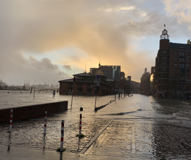 Überschwemmte Straße in Hamburg, Deutschland mit Wasser auf der Straße, Polen, Schilder, Gebäuden und einer Brücke unter einem bewölkten Himmel.