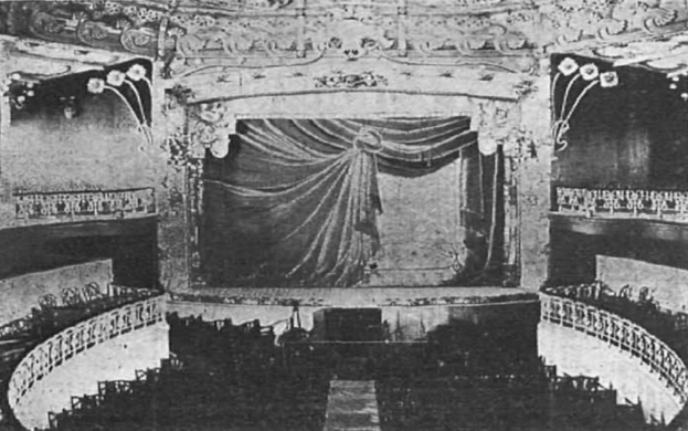 Schwarzes und weißes Foto des Auditoriums des Theatre Royal in London, das eine Bühne mit einem Vorhang, Geländern und ornamentalen Wanddesigns zeigt, mit ein paar sitzenden Menschen und Text unten.