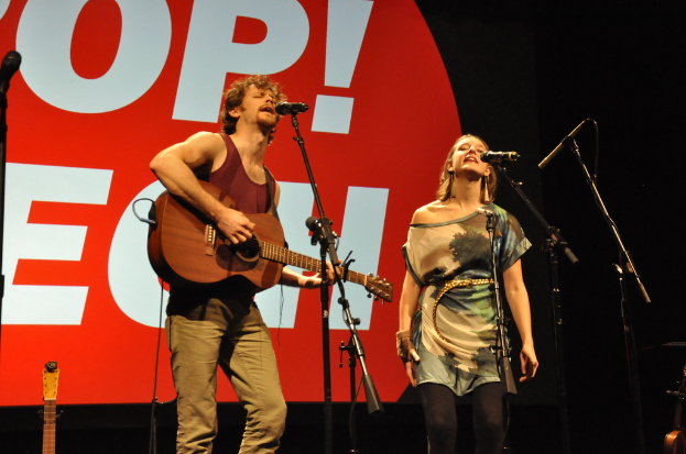 Ein Mann und eine Frau singen in Mikrofone, wobei der Mann eine Gitarre spielt und im Hintergrund ein Banner zu sehen ist.