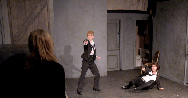 Zwei Frauen in schwarzen Anzügen, eine sitzt auf dem Boden und hält eine Waffe, die andere steht und hält eine Waffe, mit einer dritten Frau in der Nähe, in einem Raum mit einem Stuhl, einer Toilette und einer hölzernen Tür.