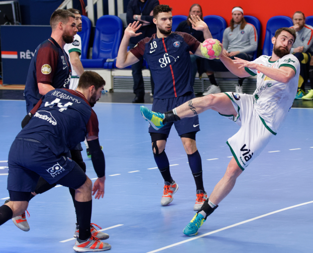 Eine Gruppe von Männern, die Handball auf einem Platz spielen, wobei einer den Ball hält und Zuschauer im Hintergrund sitzen.