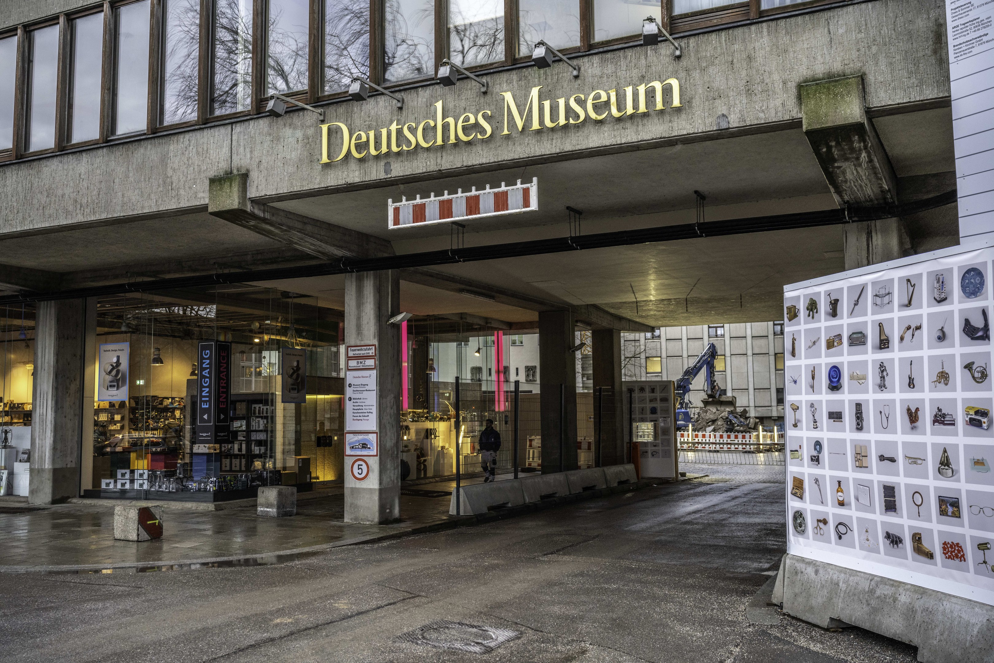 Großes Glasfrontgebäude des Deutschen Museums in Berlin mit einem Namen und einer Informationsanzeige auf der rechten Seite, vor einem Hintergrund von Bäumen und anderen Gebäuden unter dem Himmel.