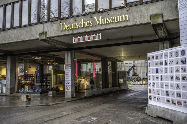 Großes Glasfrontgebäude des Deutschen Museums in Berlin mit einem Namen und einer Informationsanzeige auf der rechten Seite, vor einem Hintergrund von Bäumen und anderen Gebäuden unter dem Himmel.
