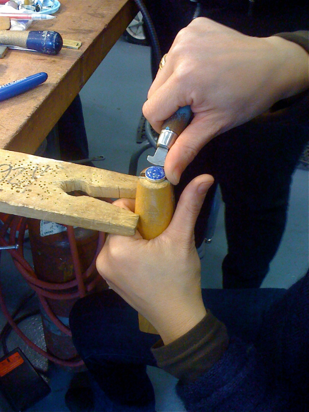 Ein Mann, der mit einem Meißel auf Holz eingraviert, sitzt an einem Tisch mit Werkzeugen, während ein anderer Mann daneben steht.