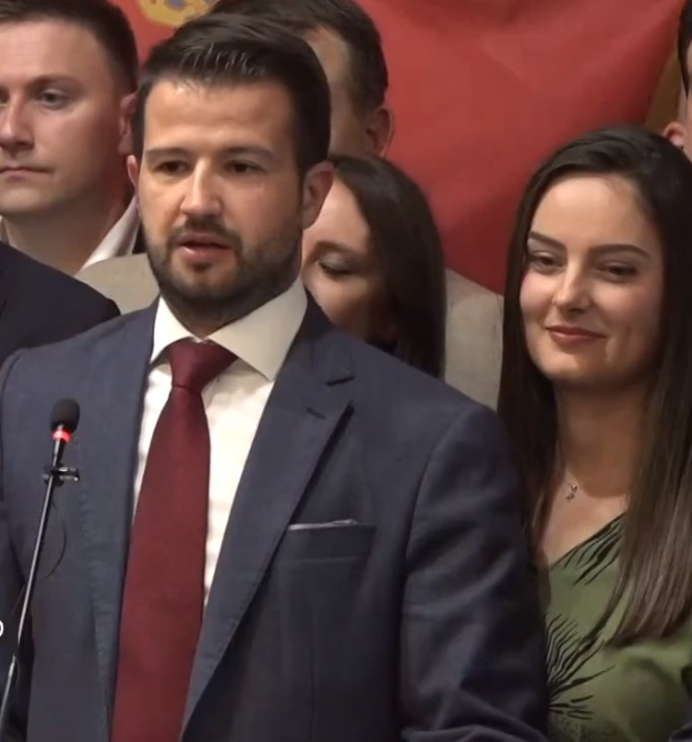 Ein Mann im Anzug spricht vor einem Publikum mit Mikrofon an einem Rednerpult, hinter einem Banner, wahrscheinlich über ein Regierungsozialmedienverbot diskutierend.