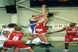 Eine Gruppe von Männern spielt Basketball auf einem Court mit einer Wand im Hintergrund, die Text und Lautsprecher zeigt, und einem Basketball in der Luft oben im Bild.