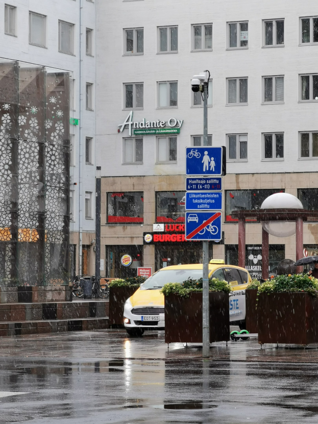 Gelbes Taxi fährt auf regennasser Stadtstraße mit einer Person mit Schirm, einer Litfaßsäule, Blumenpflanzen und einem Gebäude mit Fenstern im Hintergrund.