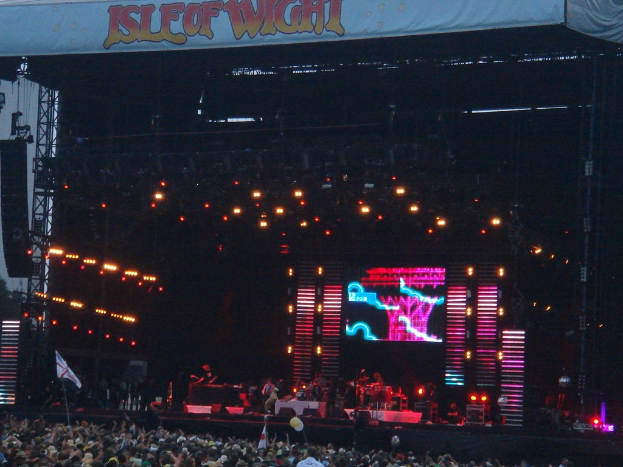 Eine Bühne mit Lichtern, einem Bildschirm und Musikern, die Instrumente spielen, mit einem Banner oben, einer Menge unten und Fahnen mit Stangen und Luftballons.