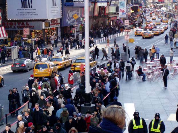 Eine belebte Straßenszene in der Stadt mit Fahrzeugen auf der Straße, Fußgängern auf den Bürgersteigen und Gebäuden mit Geschäften und Schildern auf beiden Seiten.
