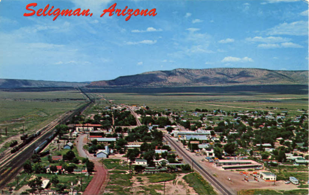 Luftaufnahme von Seligman, Arizona, mit Häusern, Bäumen, Straßen, Fahrzeugen, Pfählen, Gras, Hügeln und einem textbehafteten Himmel.