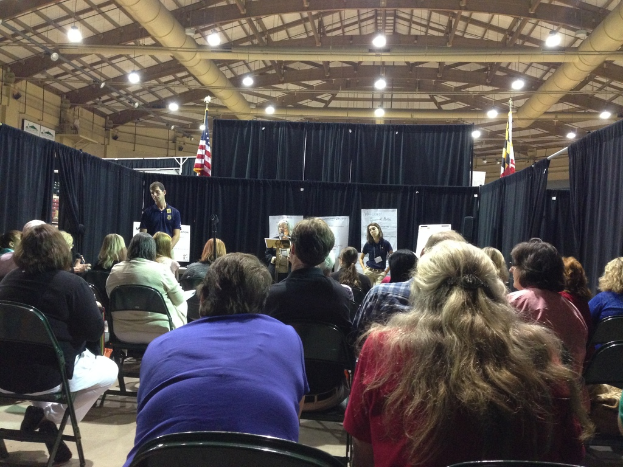 Eine Gruppe von Menschen, die auf Stühlen sitzen, mit einer Person, die an einem Podium steht, in einem Raum mit Bannern, blauen Vorhängen, Flaggen und Deckenleuchten.