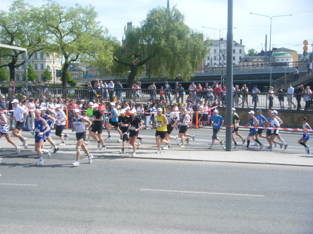 Gruppe von Menschen beim Marathon auf einer Straße mit Absperrungen, Zuschauern und Zielband unter einem bewölkten Himmel.