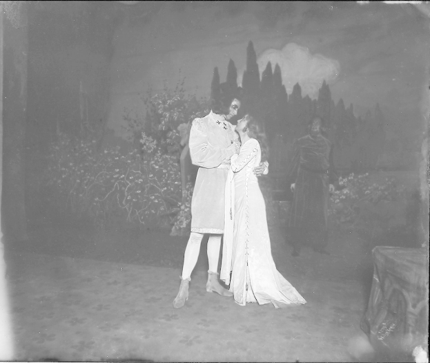 Ein Schwarz-Weiß-Foto eines Mannes und einer Frau, die sich auf einer Bühne umarmen und küssen, mit einem Publikum, Pflanzen und Bäumen im Hintergrund.