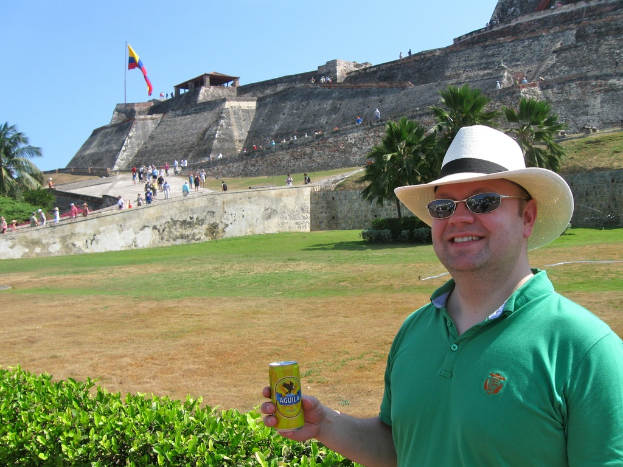Eine Person in einem grünen T-Shirt und Brille steht links, hält eine Bierdose und hat Pflanzen und ein Schloss mit Menschen und einer Fahne im Hintergrund.