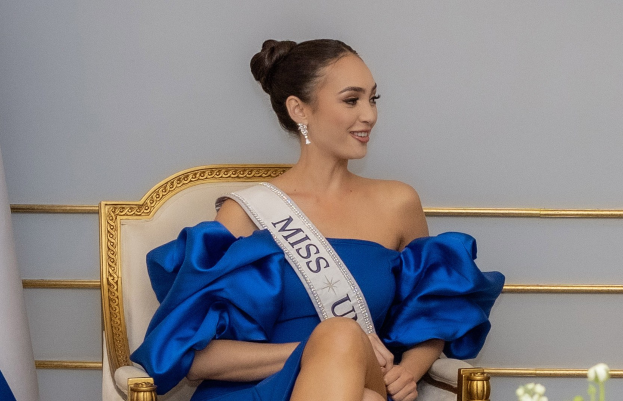 Eine Frau in einem blauen Kleid trägt einen 'Miss Universe 2019'-Schal und sitzt lächelnd auf einem Stuhl, umgeben von Blumen und einer Wand im Hintergrund.