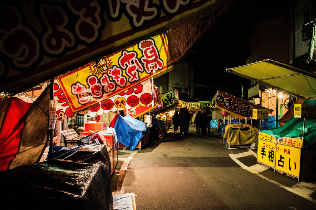 Ein belebter Nachtmarkt mit zahlreichen beleuchteten Ständen, die eine Straße säumen, Menschen, die zwischen ihnen umhergehen, und Gebäude auf beiden Seiten, mit einer Person, die im Vordergrund steht.