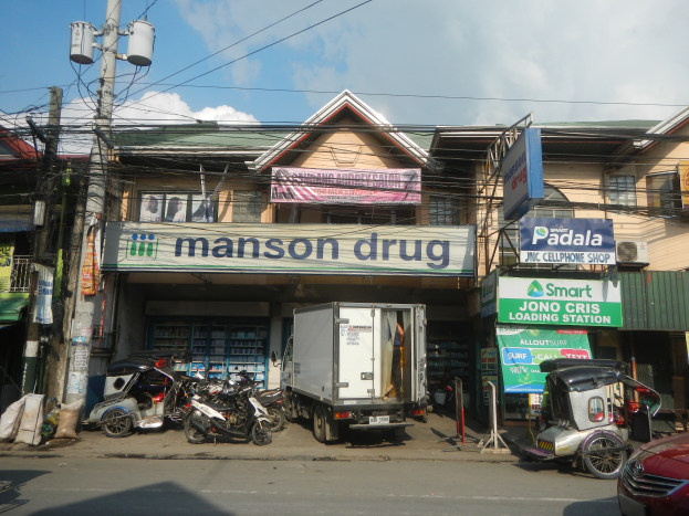 Eine Straßenszene mit einem vor einem Gebäude mit der Aufschrift "Manson Drug" geparkten Lkw, Fahrzeugen auf der Straße, Strommasten mit Drähten und Schildern, unter einem bewölkten Himmel.