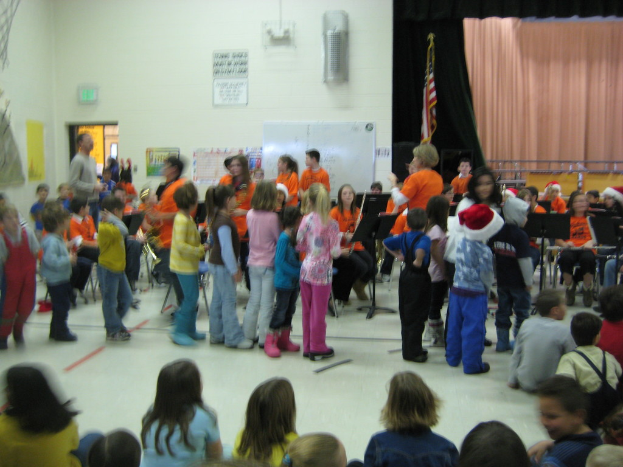 Kinder in bunten Kleidern sitzen unten im Bild, mit einer Bühne, einem Vorhang, einer weißen Tafel, Plakaten, einer Tür, Schildern, einer Flagge und einem Gegenstand an der Wand im Hintergrund.