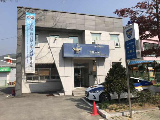 Polizeiauto vor dem Seoul Police Headquarters, einem Gebäude mit Glasfenstern und -türen, umgeben von Bäumen, Pflanzen, Verkehrskegeln und einer Fahne mit Text, mit Hügeln und einem klaren blauen Himmel im Hintergrund.