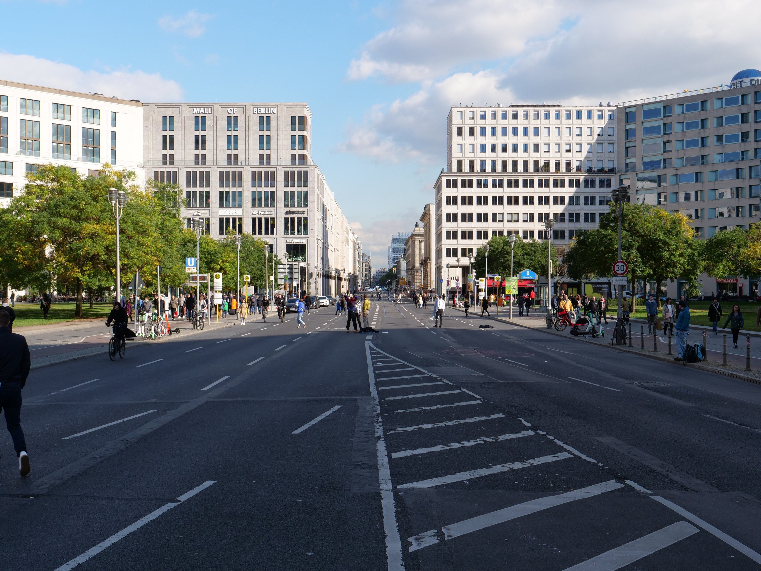 Eine belebte Stadtstraße in Berlin, Deutschland, mit Fußgängern und Radfahrern auf der Straße, flankiert von hohen Gebäuden, Bäumen, Laternenmasten und Schildern unter einem bewölkten Himmel.