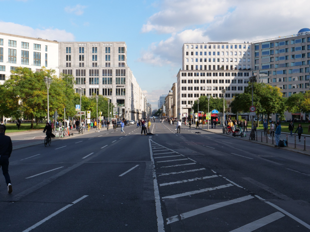 Eine belebte Stadtstraße in Berlin, Deutschland, mit Fußgängern und Radfahrern auf der Straße, flankiert von hohen Gebäuden, Bäumen, Laternenmasten und Schildern unter einem bewölkten Himmel.