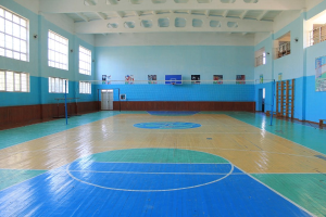 Große Turnhalle mit blau-weißem Boden, die einen Basketballplatz, ein Netz mit Stangen, eine Tür, Fenster, Wandplakate, ein Geländer und ein Dach zeigt.