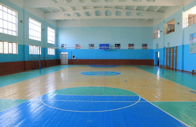 Große Turnhalle mit blau-weißem Boden, die einen Basketballplatz, ein Netz mit Stangen, eine Tür, Fenster, Wandplakate, ein Geländer und ein Dach zeigt.
