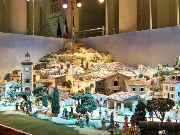 Eine Museumsausstellung eines Krippenmodells mit Menschen, Häusern, Bäumen, Felsen, einem Kirchturm, Flaggen, Pfählen und Lichtern, alles beleuchtet von Museumsbeleuchtung.