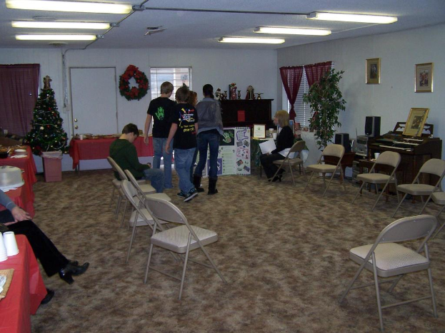 Ein Raum mit mehreren Stühlen, Tischen und Menschen, einige stehend und einige sitzend, mit Tassen und Gefäßen auf den Tischen, einem Weihnachtsbaum, einem Piano auf der rechten Seite, Fotorahmen, Fenstern, Röhrenleuchten an der Decke und einer Wand mit einem Strauß, Vorhang, Tür und weiteren Fotorahmen im Hintergrund.