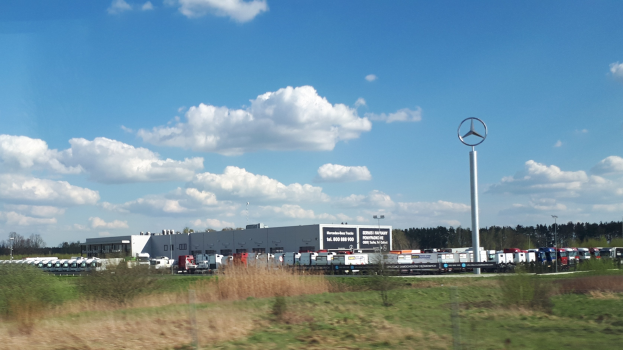 Ein Mercedes-Benz-Werk in Deutschland mit Fahrzeugen auf der Straße, umgeben von Gras, Pflanzen, Bäumen, Gebäuden und Strommasten, darunter einer mit einem Mercedes-Benz-Logo, unter einem bewölkten Himmel.