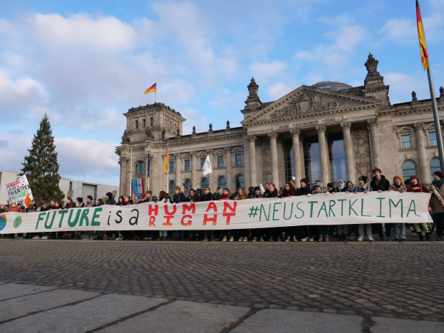 Eine Gruppe von Menschen steht vor dem Reichstagsgebäude in Berlin und hält ein Transparent mit der Aufschrift "Zukunft ist ein Menschenrecht", während das Gebäude mit Säulen, Fenstern und Bögen geschmückt ist und von Bäumen und Fahnen umgeben ist, unter einem bewölkten Himmel.