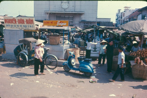 A busy city street with shops, tents, umbrellas, an auto rickshaw, a motorcycle, pedestrians, and buildings, with litter visible on the road.