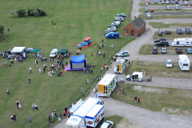 Eine Vogelperspektive eines Parkplatzes mit vielen Fahrzeugen, einigen Menschen in der Nähe von Zelten, einer zentralen Hütte und ein paar Bäumen auf der linken Seite.