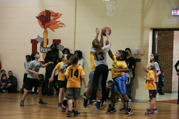 Kinder in gelben und grauen T-Shirts spielen Basketball, wobei ein Mädchen in einem grauen T-Shirt mit der Nummer 13 einen Wurf versucht. Im Hintergrund sitzt ein Mann in einem blauen Hemd in der Nähe einer weißen Wand mit Text, und rechts davon ist ein Ausgangsschild und ein rotes Backsteingebäude zu sehen.