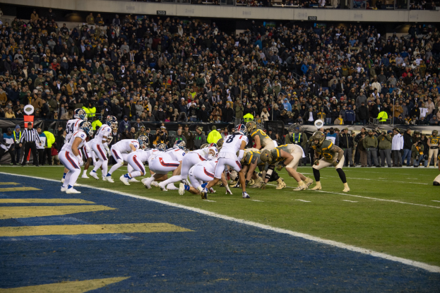 Ein Fussballspiel im Gange mit Spielern in Sportkleidung und Helmen auf dem Feld, Zuschauern, Geländern und Tafeln mit Text im Hintergrund.