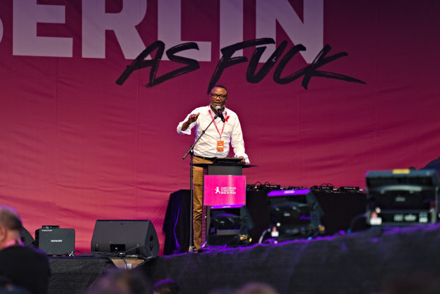 Ein Mann in einem weißen Hemd und braunen Hosen steht auf einer Bühne und hält ein Mikrofon, während er eine Menge anspricht. Im Hintergrund ist ein pinker Banner mit der Aufschrift "Berlin as Fuck" zu sehen.